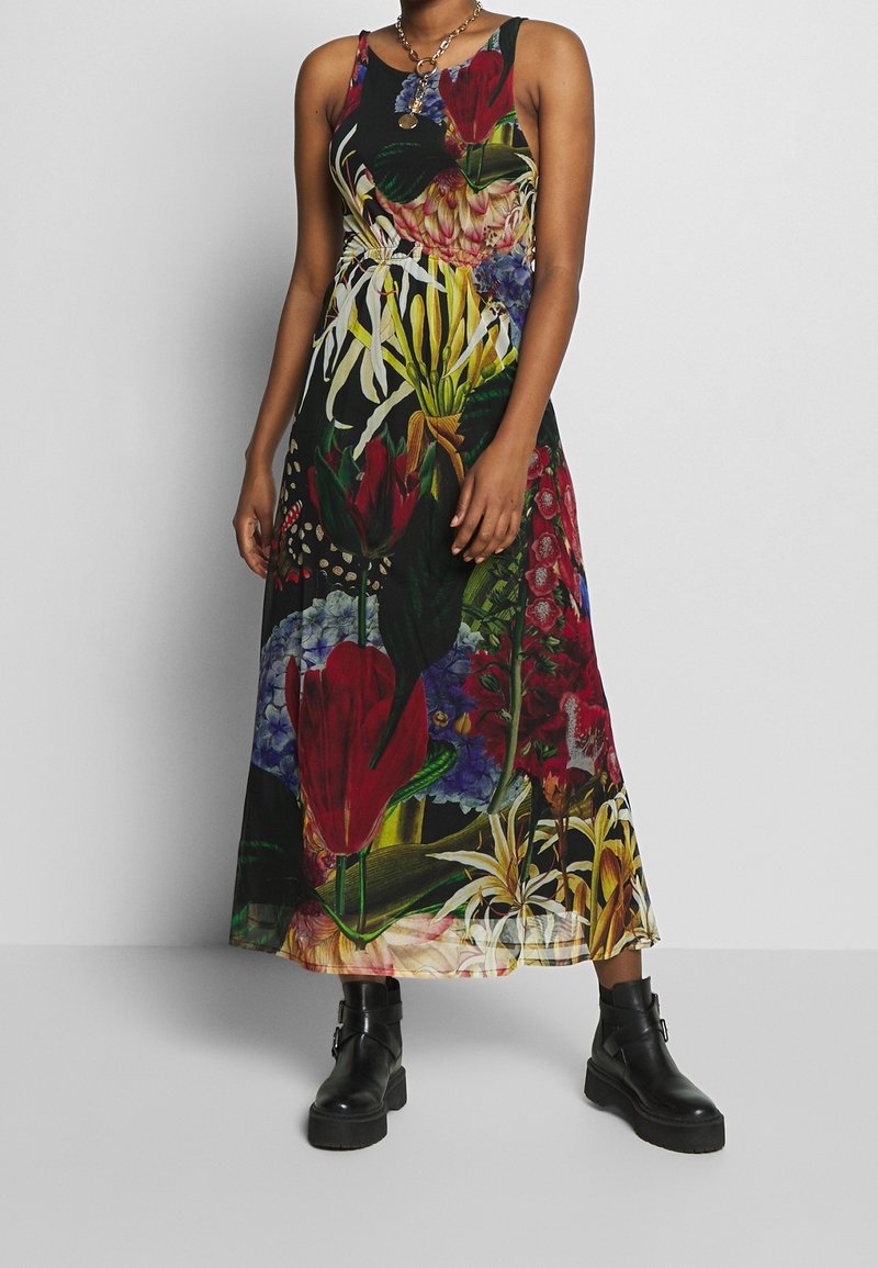 Kvinde iført en ærmeløs sort kjole med stort farverigt blomsterprint og kraftige sorte ankelstøvler, står foran en ensfarvet baggrund.