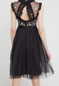 Robe noire avec corsage brodé de fleurs orné de dentelle et détails volantés aux épaules, jupe fluide en tulle. Dos nu.