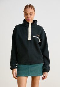 Pullover nero in pile con colletto alto e patta di bottoni color crema; presenta una tasca a zip frontale e orlo elasticizzato. Gonna teal sotto.
