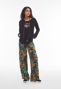 Chaqueta negra con cremallera y forro de patrón de hojas coloridas, combinada con pantalones negros holgados que presentan un vibrante estampado de hojas tropicales. Zapatillas negras.