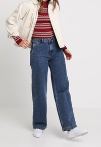 Romige ribfluwelen jas over een rood-wit gestreepte trui, gecombineerd met hoge blauwe wijde jeans en witte sneakers.