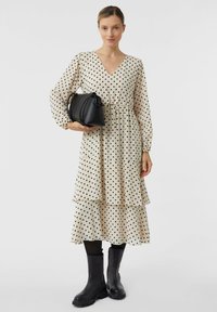 Beżowa sukienka w groszki z dekoltem w serek i warstwowym fasonem; do niej czarna torebka i czarne kozaki za kolano. Gładka, lejąca się tkanina.