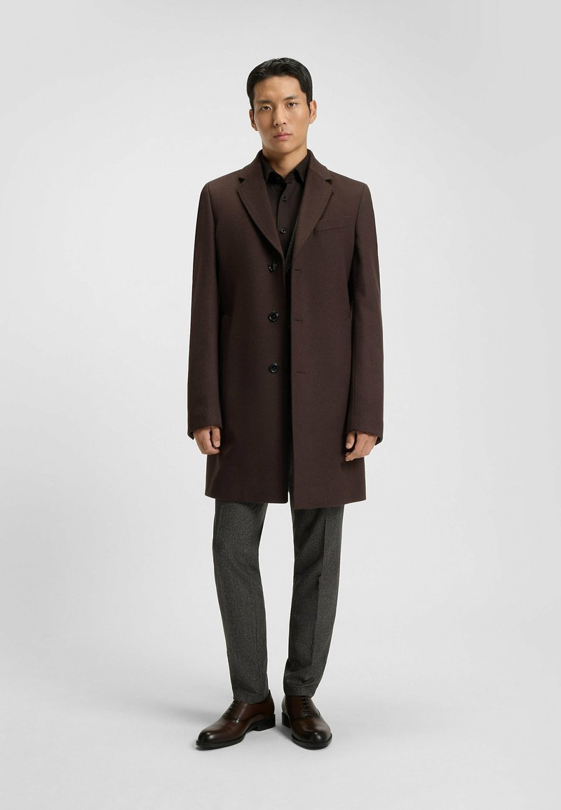 Brown wool coat with a tailored fit, single-breasted design, black buttons, and a chest pocket, paired with gray textured trousers and brown shoes.