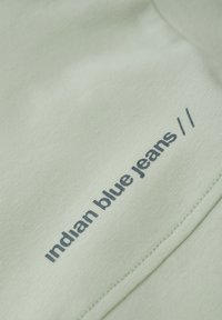 Tissu vert clair avec une surface texturée. Présente un texte marine "jeans bleu indien" et une couture décorative le long du bord.