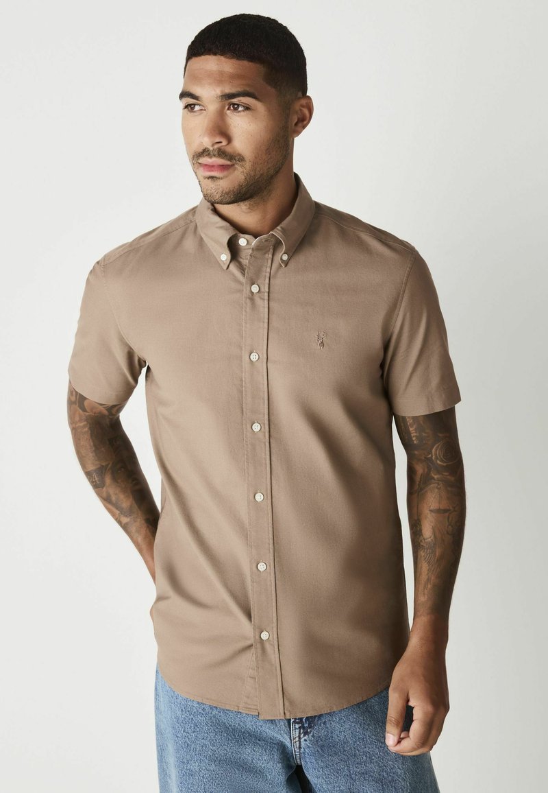 Kortærmet skjorte i beige stof med spids krave, frontknapper og logo på brystet. Kombineret med blå denim.