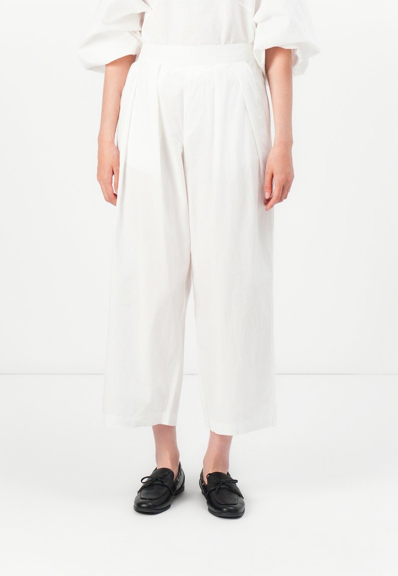 Pantaloni bianchi a gamba larga con pieghe davanti, realizzati in tessuto leggero. Abbinati a scarpe nere con lacci.