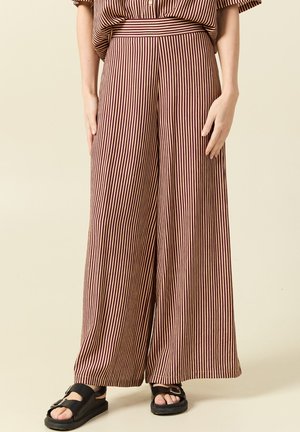 Donna che indossa pantaloni a gamba larga a vita alta con righe verticali marroni e beige, abbinati a sandali neri con fibbia.