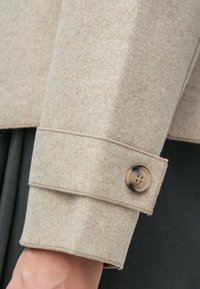 Beige erme i filt-lignende stoff med søm og mansjett. Har en rund, matt knapp med fire hull. Bakgrunn i mørkegrått.