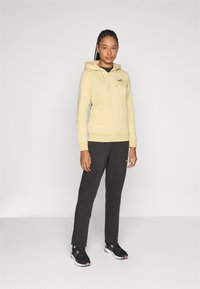 Helle gelbe Zip-Hoodie mit Kapuze, kombiniert mit dunkelorangen Jogginghosen und schwarzen Sportschuhen. Minimalistisches Design mit Logo-Akzenten.