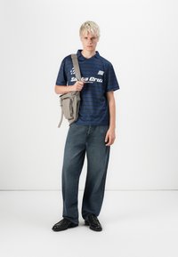Marine-stripet kortermet polo skjorte med "Santa Cruz" logo, parret med løse blå denimjeans og en grå crossover-veske. Svarte sko.