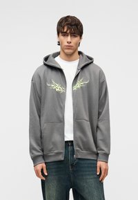 Γκρι φούτερ με φερμουάρ και κουκούλα, με κιτρινοπράσινα γραφικά στο στήθος. Μαλακό ύφασμα, τσέπες τύπου καγκουρό και ριμπ μανσέτες.