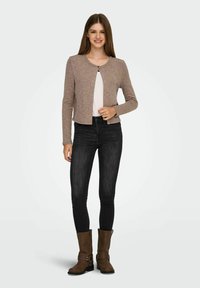 Cárdigan de punto marrón con cuello redondo, textura acanalada y cierre frontal oculto, combinado con jeans skinny negros y botas de tobillo marrones.