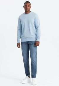 Sudadera azul claro en un tejido de punto suave, con un escote redondo y un pequeño logo en el pecho, combinada con jeans de mezclilla oscura y zapatillas blancas.