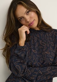 Tmavomodrá blúzka s motívom paisley v medenej farbe, so zúženým golierikom, dlhými rukávmi a textúrovanou látkou. Manžety sú zvýraznené detailom s gombíkom.