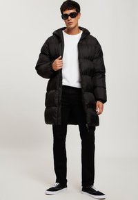 Czarna ocieplana kurtka z kapturem, z zamkiem z przodu, elastycznymi mankietami i oversize'owym krojem. Nosi ją osoba w białej koszulce i czarnych dżinsach.