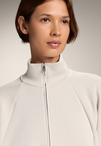 Valkoinen vetoketjullinen takki, jossa on korkea kaulus, ribbitekstuuri ja vartalonmyötäinen muotoilu, jota korostavat hopeinen vetoketju ja sauman yksityiskohdat.