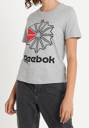 Persoon draagt een grijs Reebok T-shirt met een zwart-rood sterburst-logo, gecombineerd met zwarte spijkerbroek.