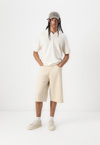 Kremfarget strikket polo-skjorte med krage, beige shorts og grå caps. Lyse sneakers og hvite sokker fullfører antrekket.