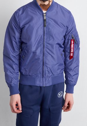 Mann trägt eine blaue Bomberjacke mit gerippten Bündchen und einem roten "Remove Before Flight"-Anhänger an der linken Ärmeltasche sowie dunkelblaue Nike Air-Hose.