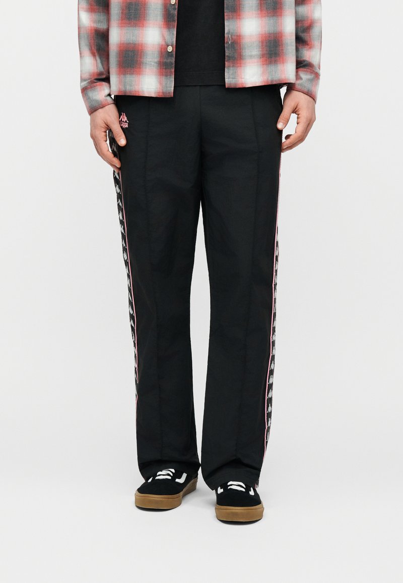 Άνδρας που φοράει μαύρο φόρμα με λευκές και ροζ λωρίδες λογότυπου στα πλαϊνά, συνδυασμένη με μαύρα αθλητικά παπούτσια με λευκά κορδόνια και καφέ σόλες.