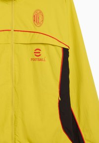 Gul jakke med lynlås foran, røde detaljer og sorte sidelapper. Har logoer: ACM 1899 og FODBOLD. Letvægts, tekstureret stof.