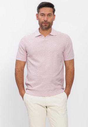 Man staat met handen in zijn zakken, draagt een lichtroze poloshirt met korte mouwen en crèmekleurige broek tegen een witte achtergrond.