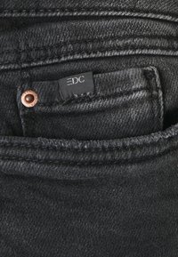 Jeans en denim noir avec un rivet en cuivre et une étiquette en tissu noir portant la mention "EDC" visible dans la poche avant. Tissu texturé.