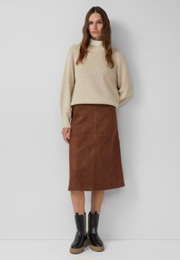 Femme portant un pull en tricot beige sur un col roulé blanc, une jupe en daim marron jusqu'aux genoux, et des bottes noires épaisses, se tenant contre un mur uni.
