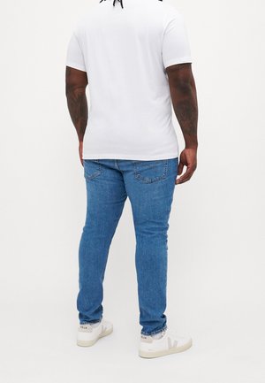 Uomo con tatuaggi che indossa una maglietta bianca, jeans blu e sneakers bianche Veja, in piedi di spalle su uno sfondo semplice.