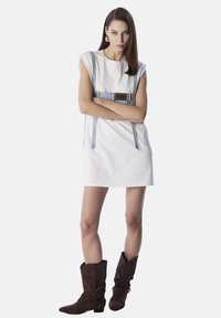 Vestido blanco sin mangas con tirantes de denim y un parche rectangular con el logo. La modelo lo combinó con botas altas de ante marrón. Diseño minimalista.