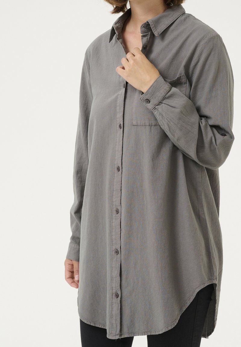Personne portant une chemise grise ample à manches longues avec poche avant, tenant le col, vêtue d’un pantalon noir sur fond uni.
