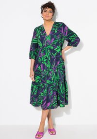 Blumengemustertes, V-Ausschnitt-Kleid in dunklem Navy mit grünen und lila Blättern, ausgestattet mit mehrlagigem Rock und halblangen Ärmeln. Kombiniert mit lila Sandalen.