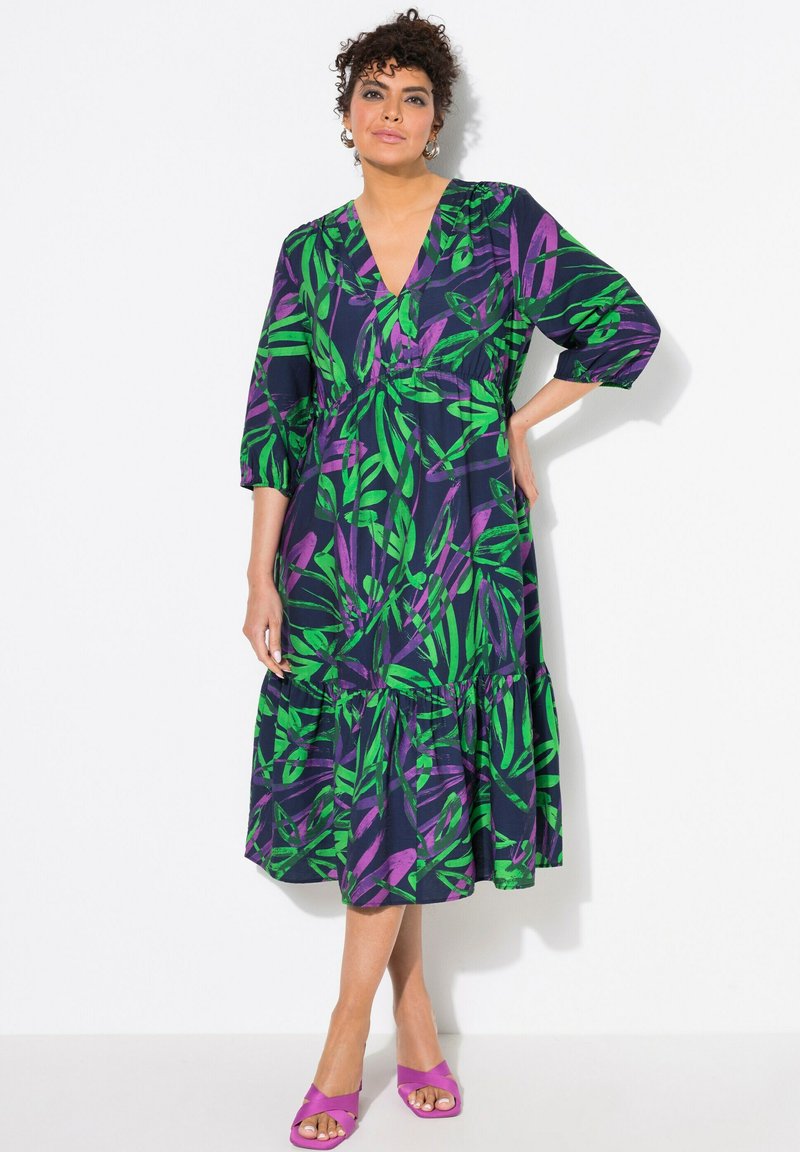 Blumengemustertes, V-Ausschnitt-Kleid in dunklem Navy mit grünen und lila Blättern, ausgestattet mit mehrlagigem Rock und halblangen Ärmeln. Kombiniert mit lila Sandalen.