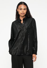 Camicia nera con bottoni in tessuto lucido e testurizzato, con taschino sul petto e maniche lunghe. Il modello indossa una vestibilità ampia, abbinata a pantaloni neri.