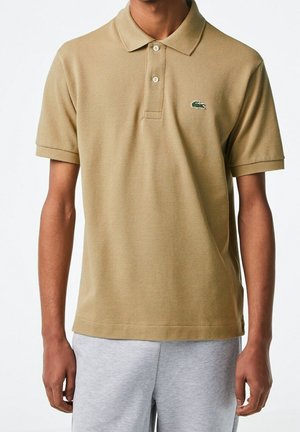 Man wearing khaki polo shirt with green crocodile logo on chest and light gray sweatpants, standing against white background.