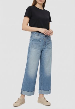 Mujer con camiseta negra de manga corta, jeans anchos de pierna con dobladillos remangados y zapatillas beige, de pie frente a un fondo liso.