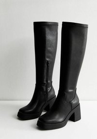 Botas negras de altura de rodilla en suave piel sintética con un tacón cuadrado, cremallera lateral y detalles de punta redondeada. Diseño limpio y elegante.