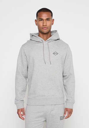 Junger Mann trägt hellgrauen Hoodie und passende Jogginghose mit minimalen schwarzen Textlogos, steht vor einem schlichten weißen Hintergrund.