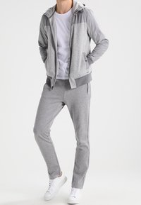 Šedá mikina s kapucí na zip a textura těla, hladké rukávy, v kombinaci se šedými tepláky. Bílí tenisky doplňují celý outfit.