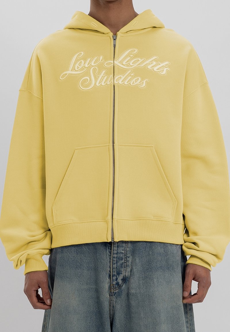 Personne portant un sweat à capuche jaune zippé avec le texte « Low Lights Studios » et un jean bleu ample, debout devant un fond uni.