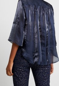 Blouse transparente bleu marine à manches courtes, texture douce, avec un détail froncé à l'arrière, associée à un pantalon foncé à motif léopard.