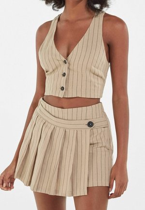 Vrouw draagt een beige mouwloze crop top met fijne strepen en knopen en een bijpassende geplooide minirok met een knoopdetail aan de zijkant.