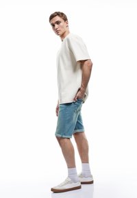 Joven con camiseta beige, pantalones cortos de mezclilla, calcetines blancos y zapatillas blancas, de pie con una mano en el bolsillo contra un fondo blanco.