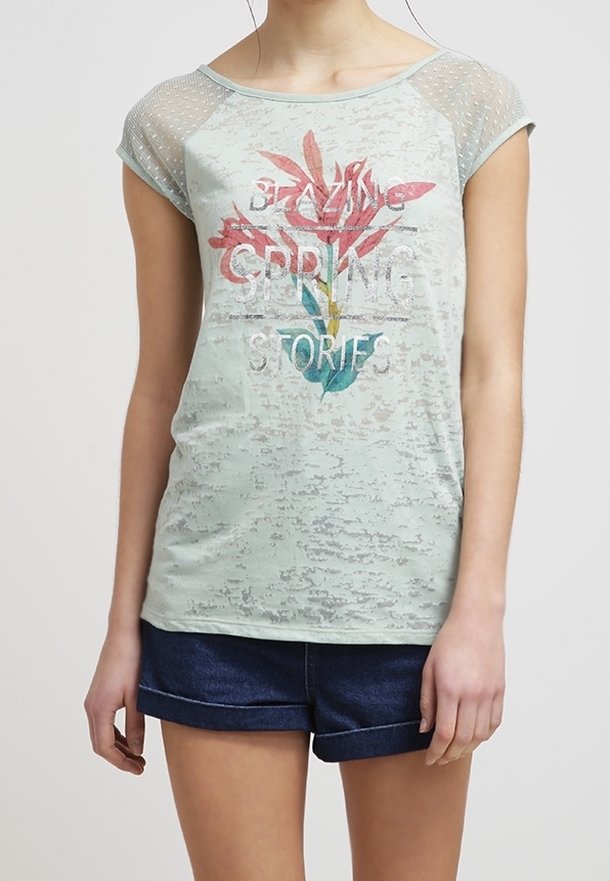 Camiseta corta de manga corta en verde claro con detalles de malla, gráfico floral y el texto "HISTORIAS DE PRIMAVERA". Combinada con pantalones cortos de mezclilla azul marino.
