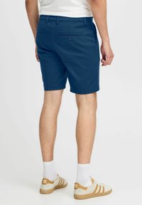 Korte marineblå shortser laget av bomullsblanding, med to baklommer med knapper, akkompagnert av hvite sokker og brune sneakers.