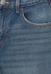 Jeans en denim bleu avec un lavage moyen, une décoloration subtile, des coutures orange et un bouton en métal avec une rivet sur la taille.