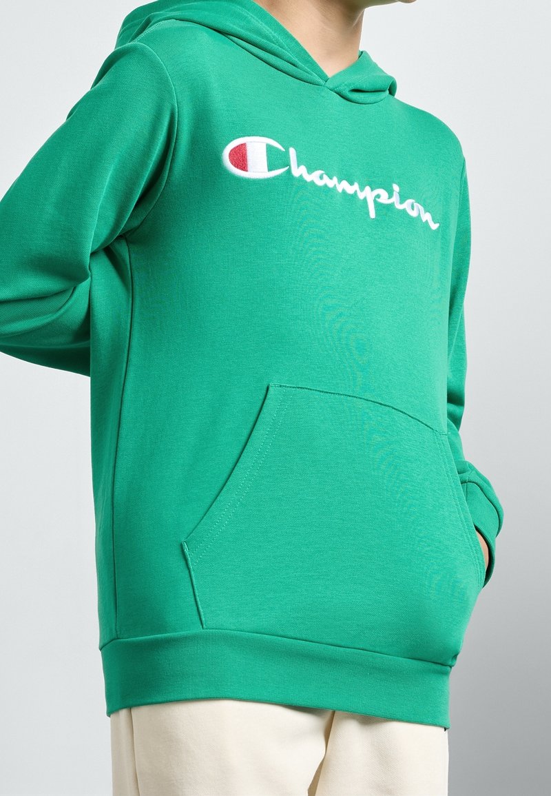 Personne portant un sweat à capuche vert Champion avec un logo blanc et rouge, mains dans la poche avant, montrée du cou aux hanches.
