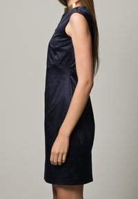 Mörkblå ärmlös klänning i en slät, glänsande tyg. Har en figurnära silhuett och vinklade sömmar för en strukturerad design.