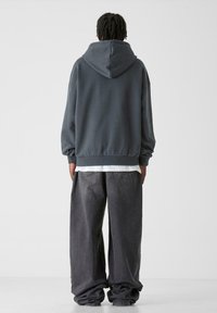 Felpa grigia oversize con cappuccio regolabile, abbinata a pantaloni in denim grigio scuro a vestibilità ampia, che mostrano una silhouette rilassata e una texture morbida.