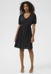 Robe noire à manches courtes avec un décolleté en V, fermeture à boutons et ceinture à la taille réglable. Texture lisse, longueur genoux, avec une jupe fluide.